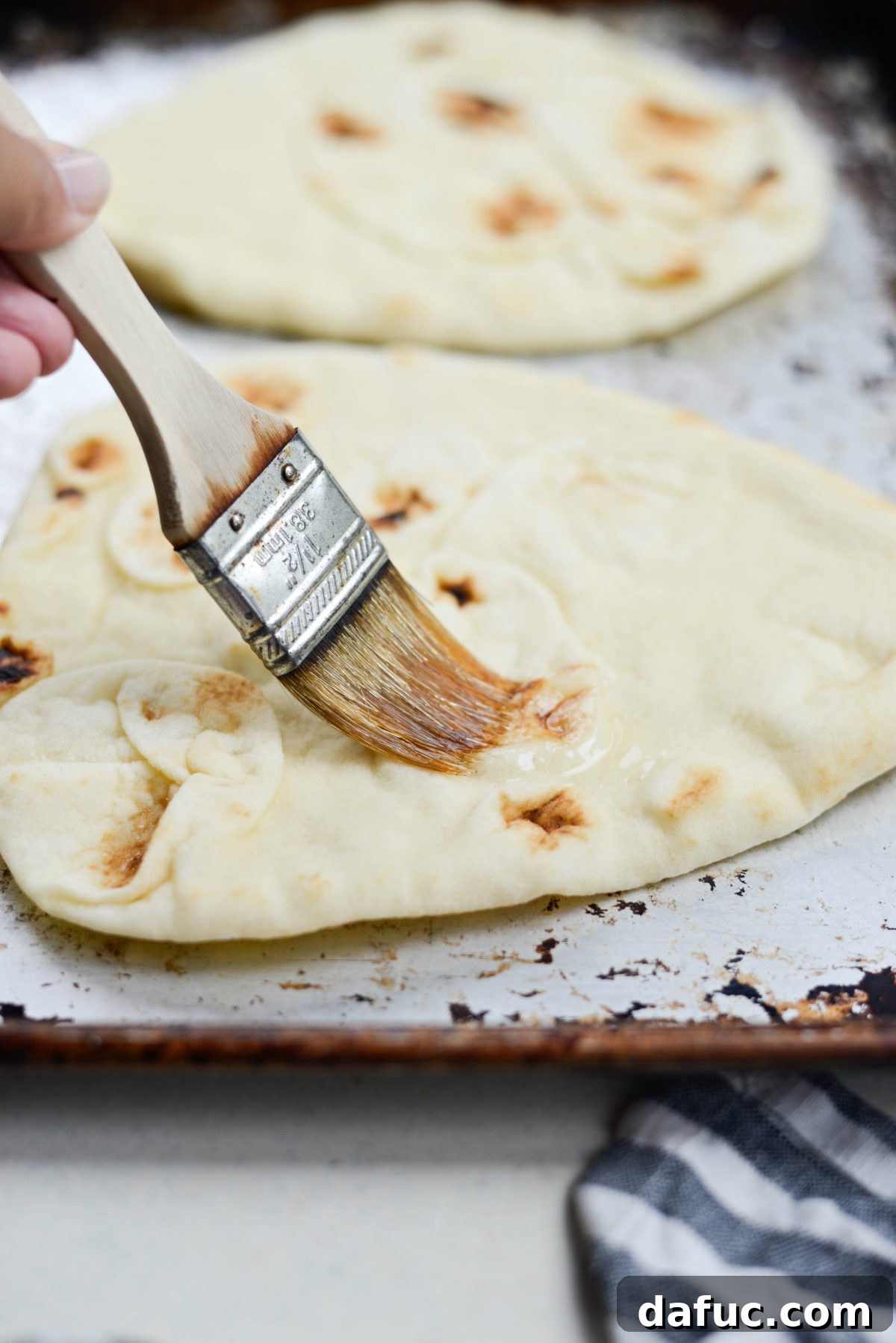 A hand lightly brushing naan bread with olive oil on a baking sheet.