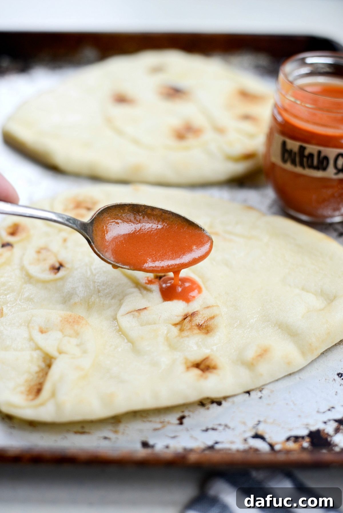 Spreading spoonfuls of homemade buffalo sauce evenly over naan bread.