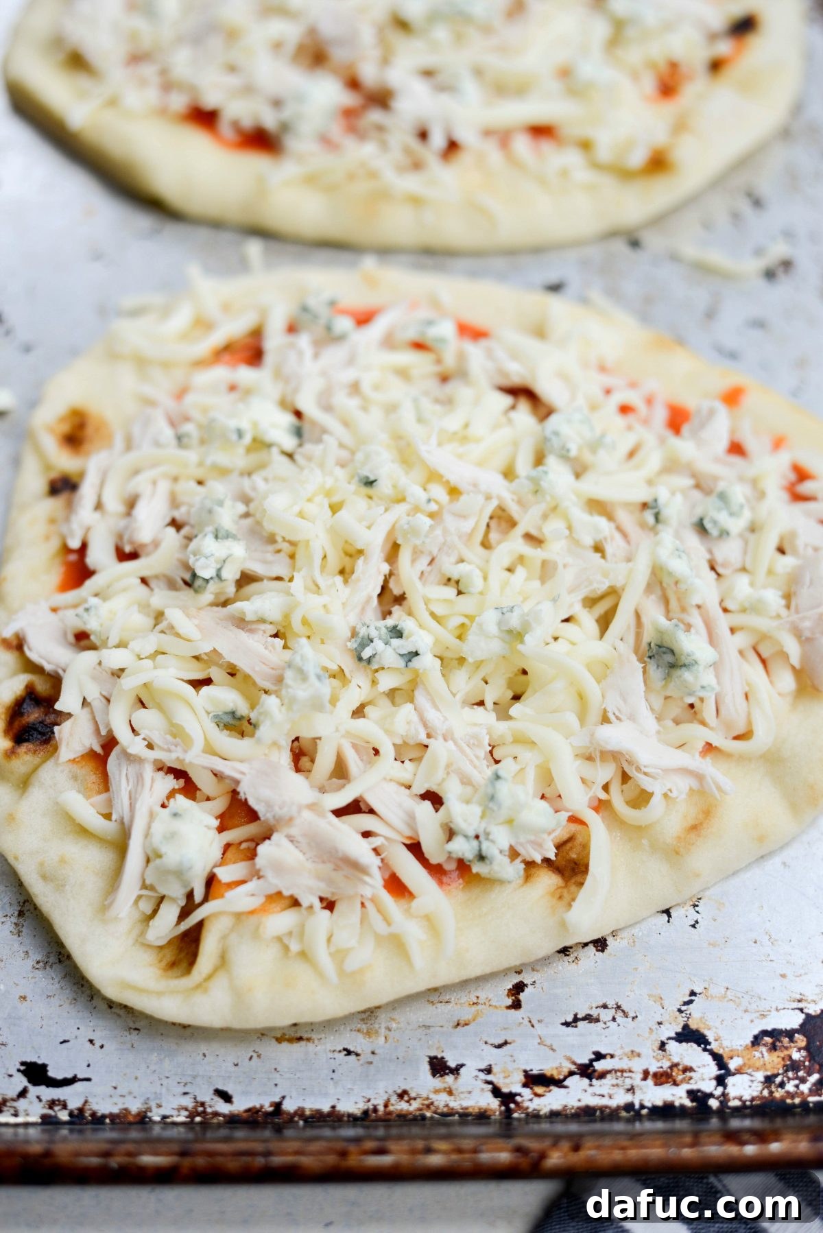 Naan pizza base topped with buffalo sauce, shredded cheese, and cooked chicken.