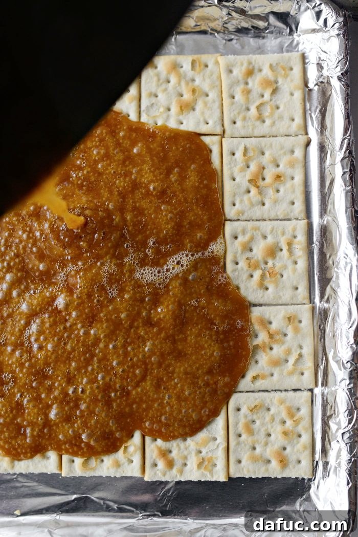 Hot butter and dark brown sugar toffee mixture being carefully and evenly poured over the arranged saltine crackers on a foil-lined baking sheet.