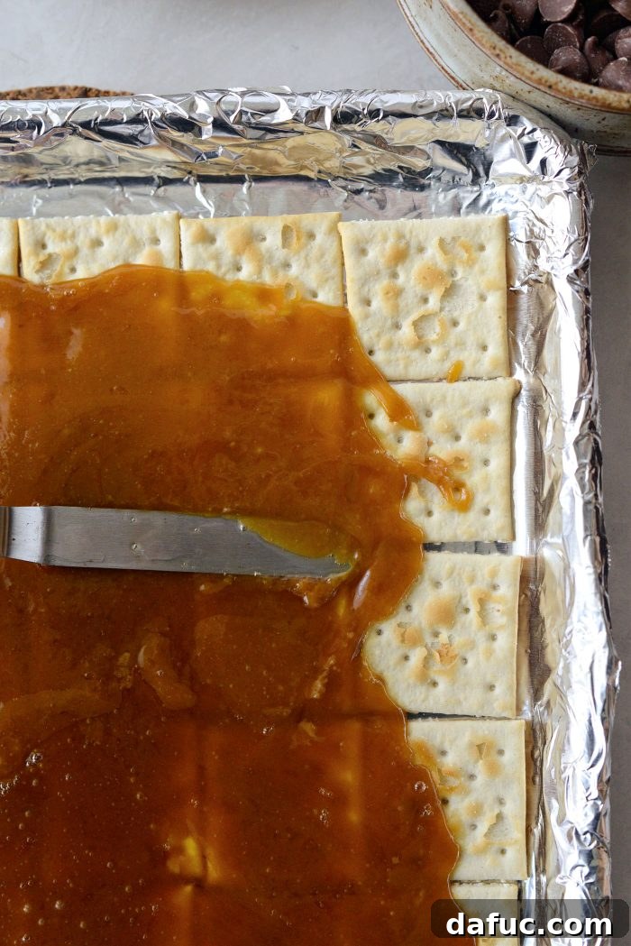 The hot toffee mixture being meticulously spread evenly over the entire surface of the saltine crackers using an offset spatula.