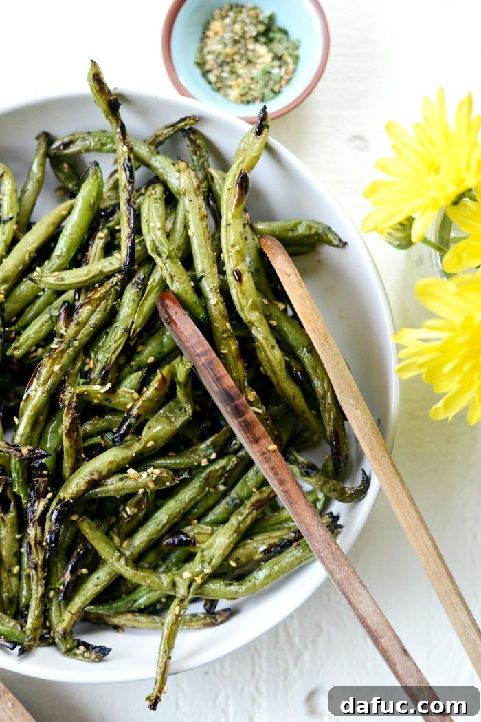 Close-up shot of tongs picking up a handful of perfectly grilled green beans from a bowl, highlighting their charred texture.