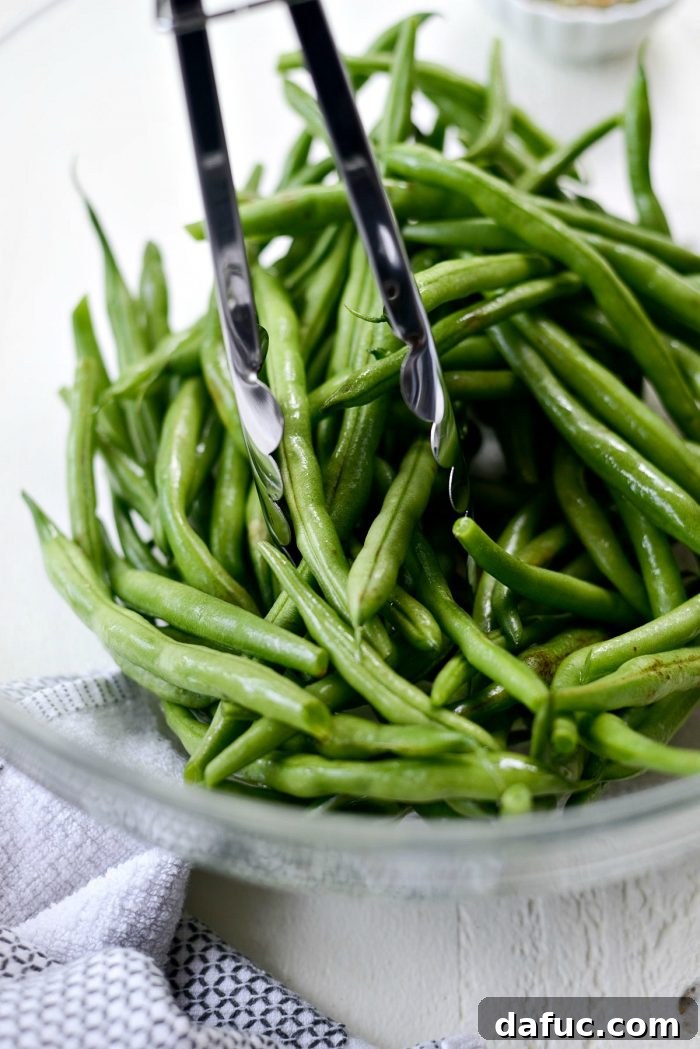 Fresh green beans being tossed with olive oil in a large glass bowl, preparing them for the grill.