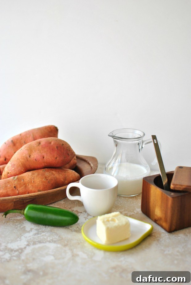 A flat lay photograph showcasing all the fresh ingredients needed for Sweet Potato Jalapeño Mash: vibrant sweet potatoes, a green jalapeño, golden butter, amber maple syrup, heavy cream, and kosher salt.