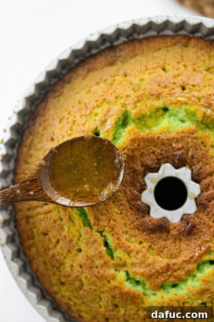 Spoonfuls of warm lemon-lime glaze being poured over the poked holes of a warm bundt cake, allowing deep absorption.