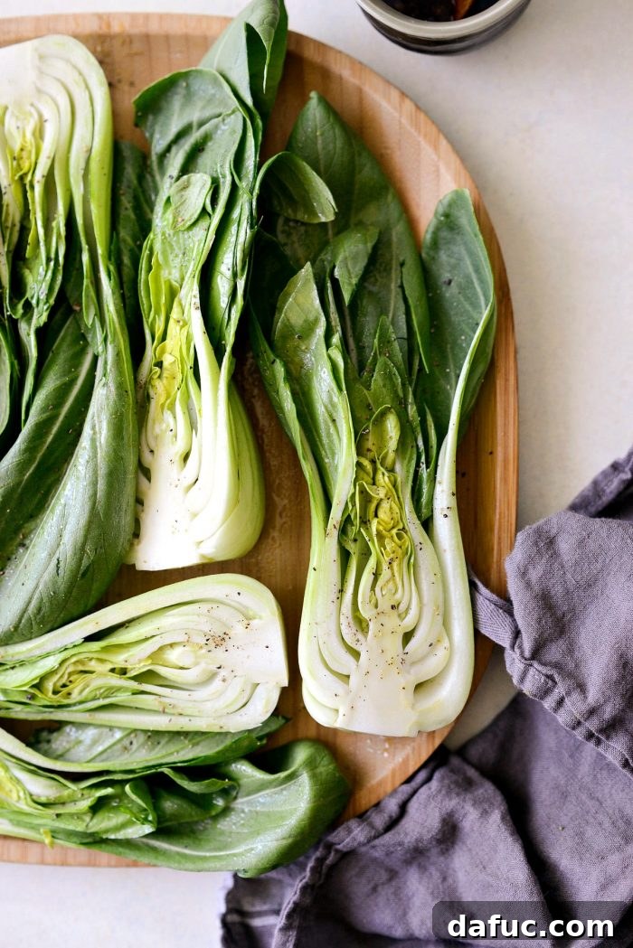 Halved baby bok choy misted evenly with olive oil and generously sprinkled with freshly ground black pepper on a cutting board