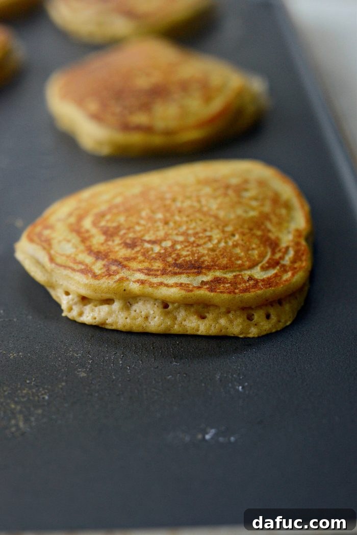 Spiced Brown Butter Pumpkin Pancakes 13 Multiple brown butter pumpkin pancakes sizzling on a hot griddle, with visible bubbles indicating they are ready to be flipped.