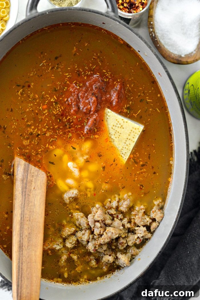 Pouring chicken broth, crushed tomatoes, white beans, and browned sausage into the pot with a parmesan rind