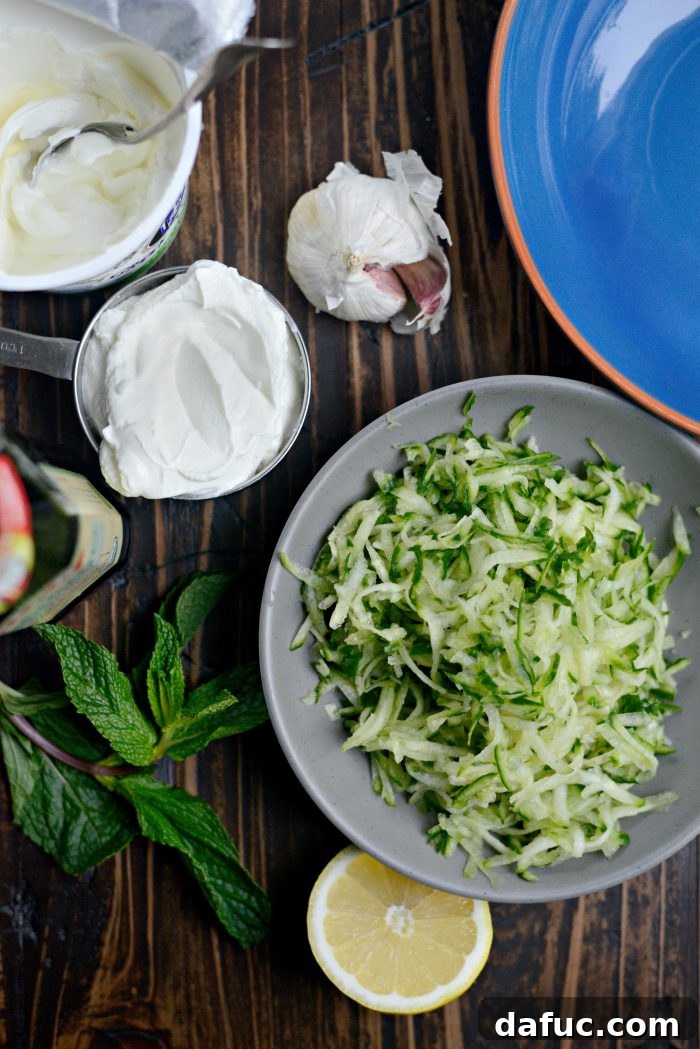 Creamy Homemade Tzatziki 3 Homemade Tzatziki ingredients - A flat lay showcasing the fresh ingredients for tzatziki: a whole English cucumber, a bowl of Greek yogurt, lemons, fresh mint, garlic cloves, and a bottle of olive oil.