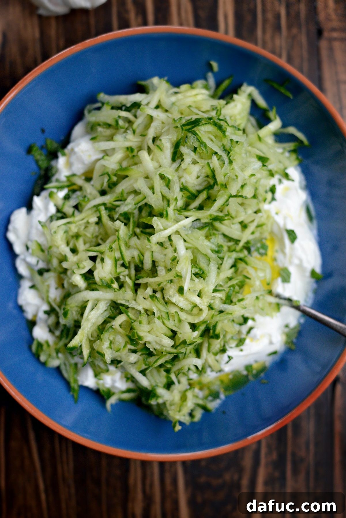 Creamy Homemade Tzatziki 5 Add grated fresh garlic and shredded and drained cucumber - Hands adding finely grated garlic and the squeezed, drained cucumber shreds into a bowl of prepared yogurt mixture.