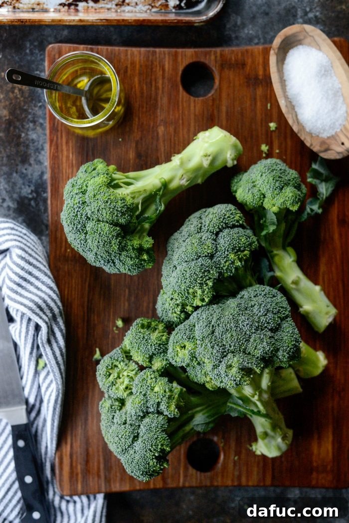 Ingredients for The Best 10-Minute Roasted Broccoli Recipe - Fresh broccoli, a bottle of olive oil, and a small bowl of kosher salt on a wooden surface.