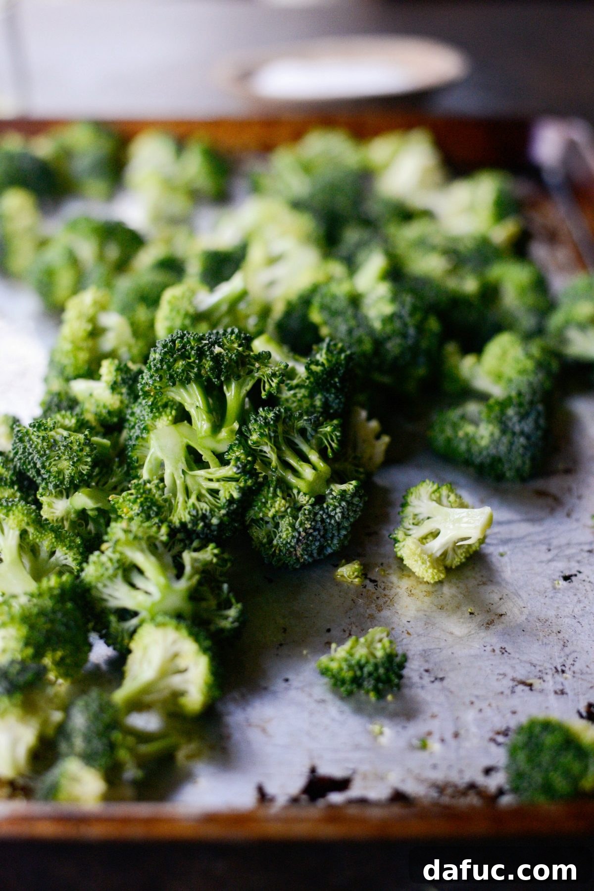 Hands tossing broccoli florets with olive oil on a baking sheet to ensure even coating.