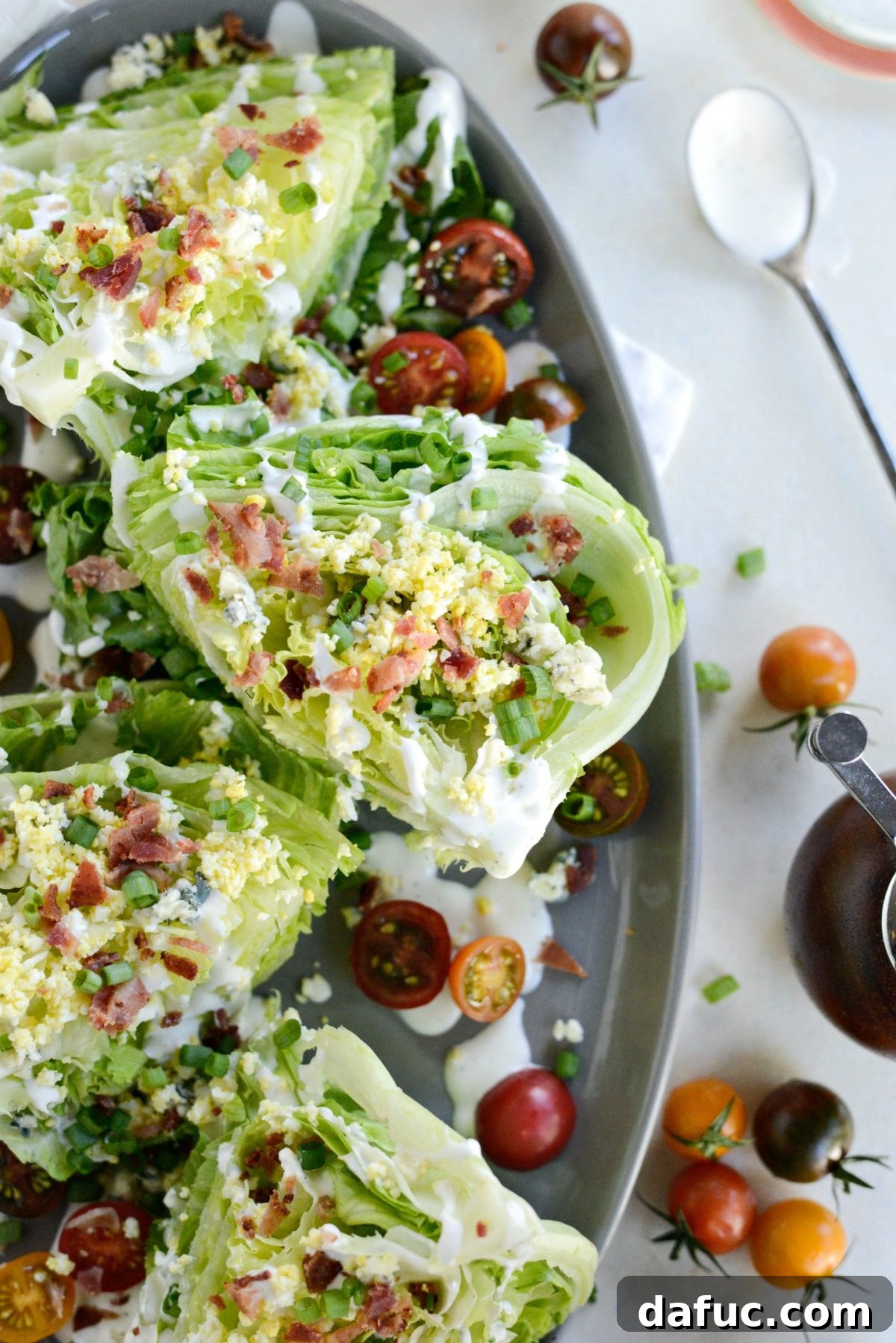 Our Signature Loaded Wedge, Drenched in Peppercorn Buttermilk 2 A vibrant Loaded Wedge Salad with Black Pepper Buttermilk Dressing, topped with crispy bacon, blue cheese, hardboiled egg, tomatoes, and green onions, presented on a white plate.
