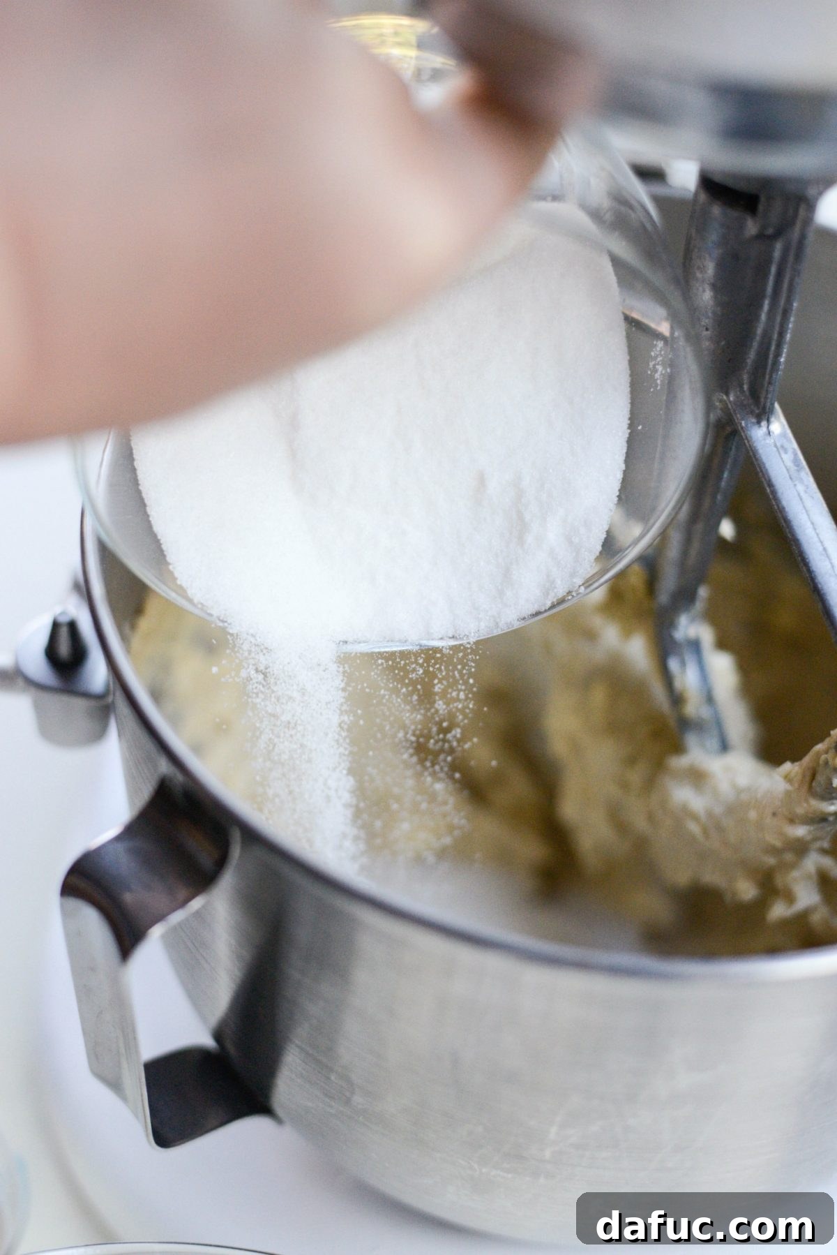 Add granulated sugar to the butter and tahini mixture - A stream of granulated sugar is being poured into the mixing bowl, joining the creamed butter and tahini, preparing for further blending.