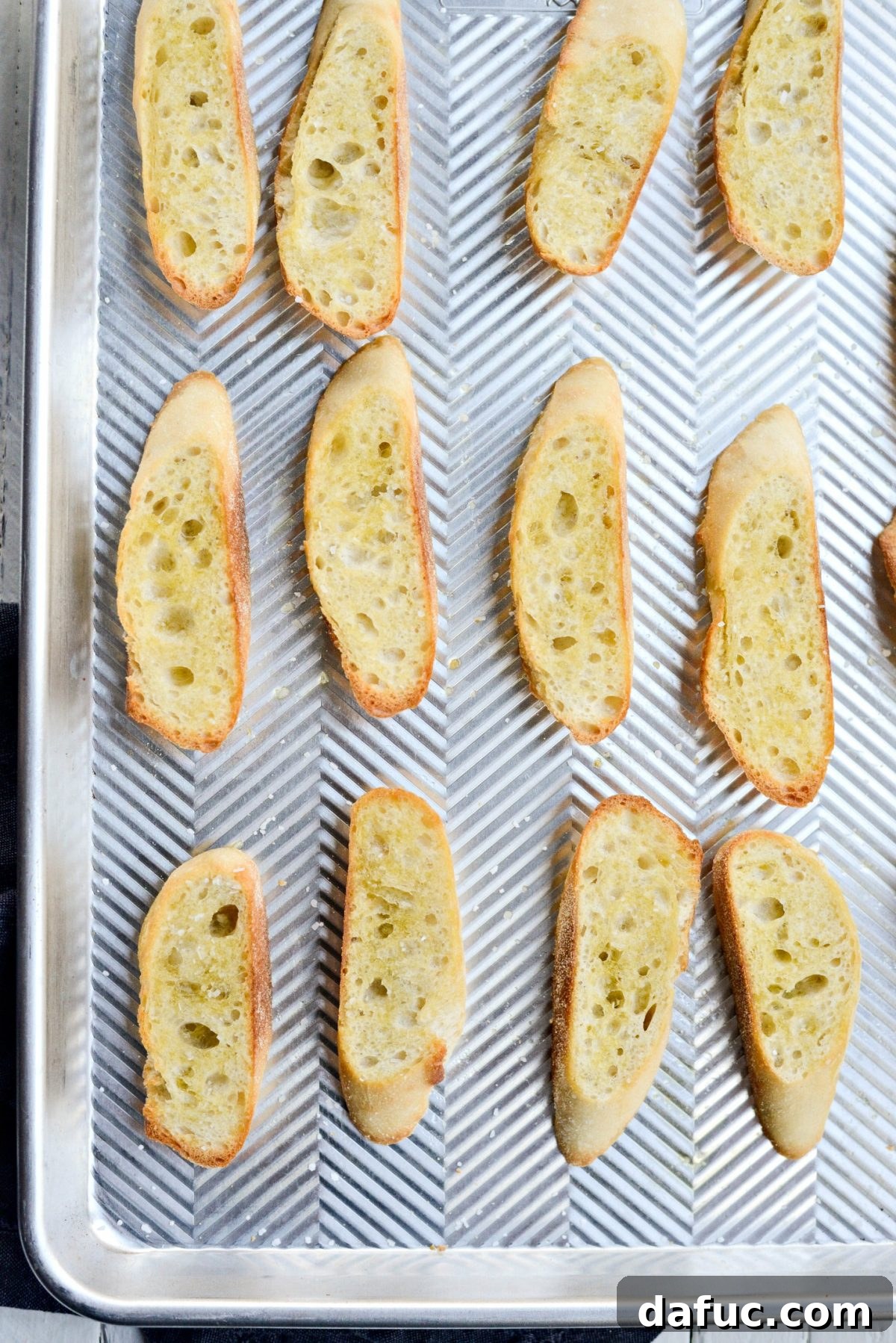 Sliced demi baguette brushed with olive oil and sprinkled with kosher salt on a baking sheet, ready for toasting.