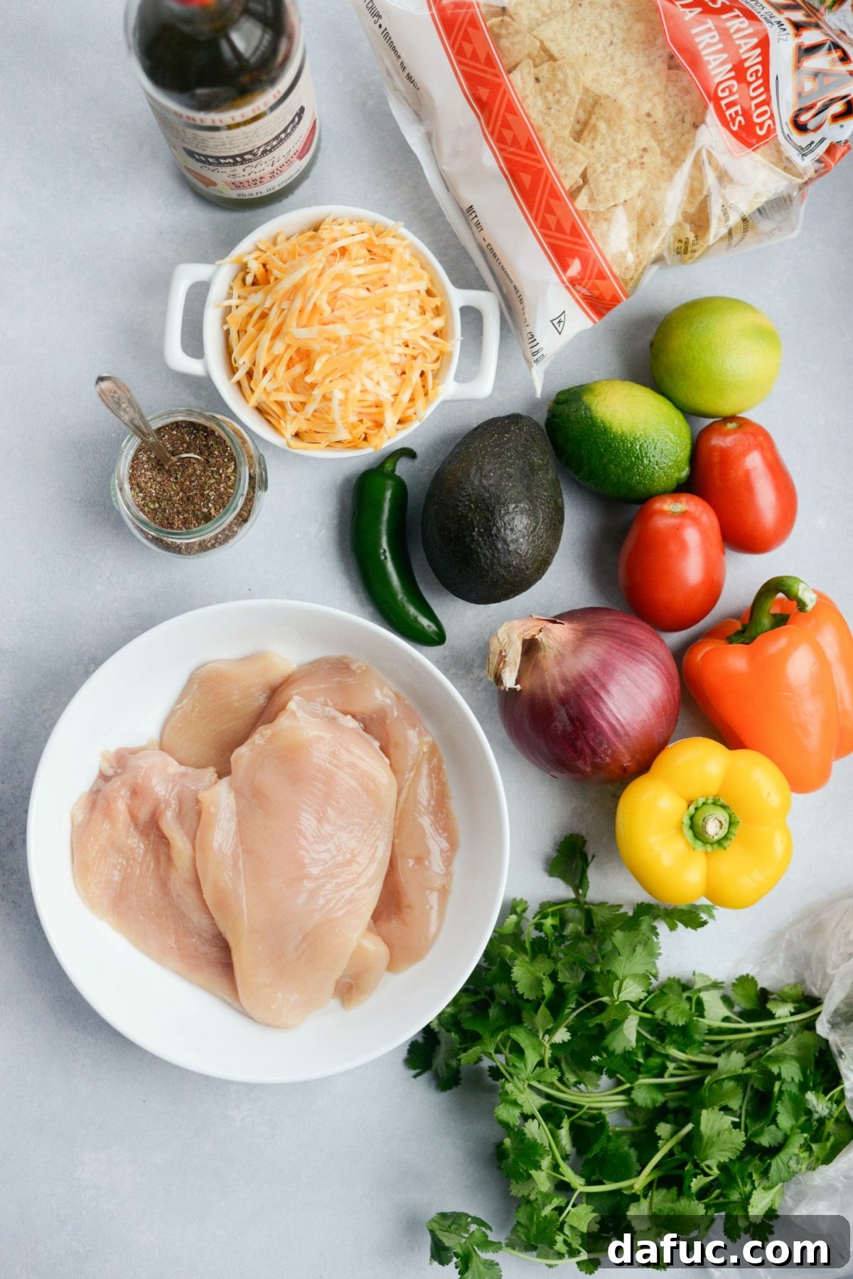 A collection of fresh ingredients for Mexicali Chicken Sheet Pan Nachos, including chicken breast, limes, bell peppers, onions, tomatoes, avocado, and tortilla chips.