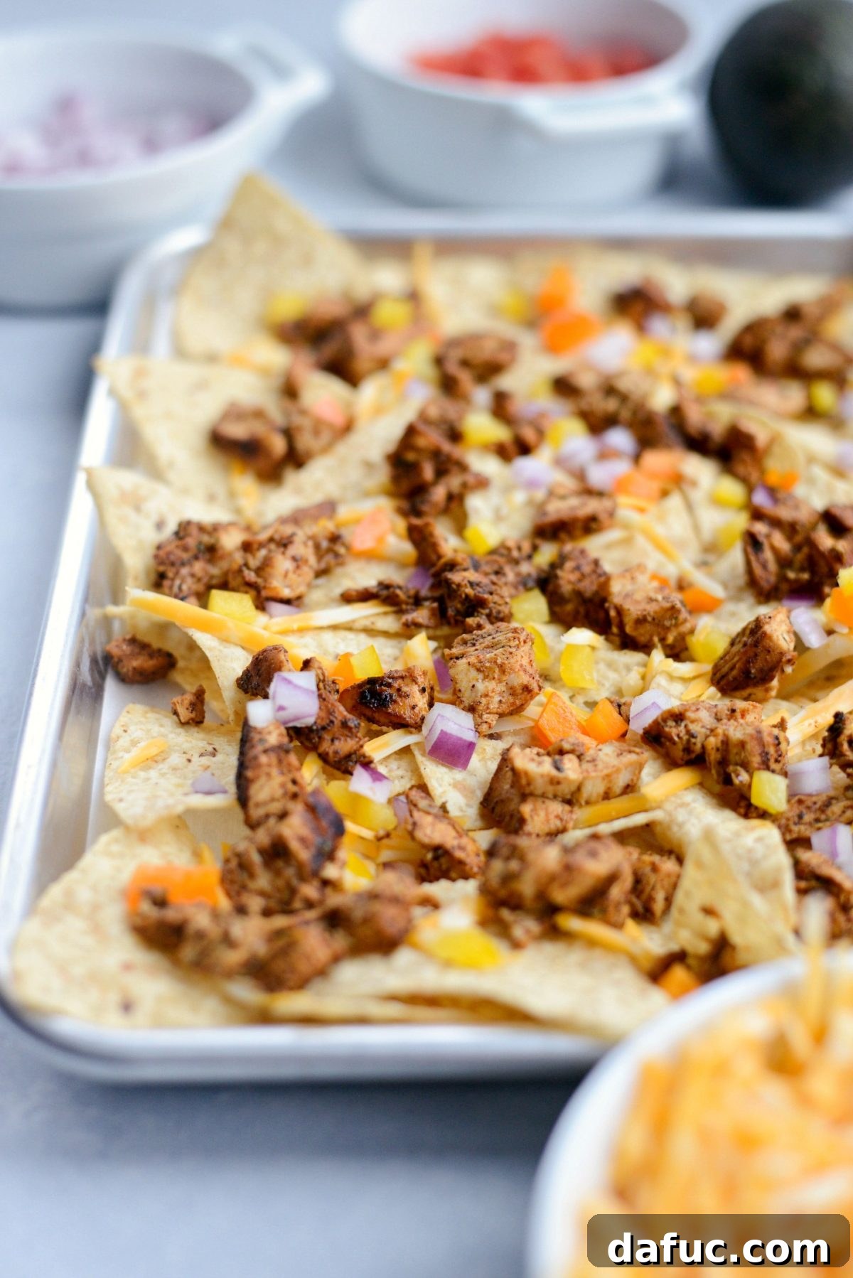 First layer of tortilla chips on a sheet pan topped with a sprinkle of shredded cheese, diced onions, bell peppers, and some cooked Mexicali chicken.