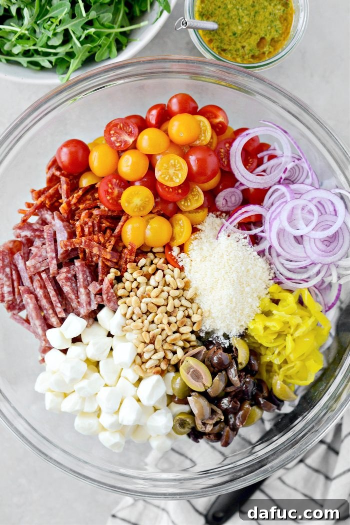 Mixing bowl filled with cooked tortellini, tomatoes, onion, pepperoncini, olives, pearl mozzarella, salami, pepperoni, toasted pine nuts, and parmesan