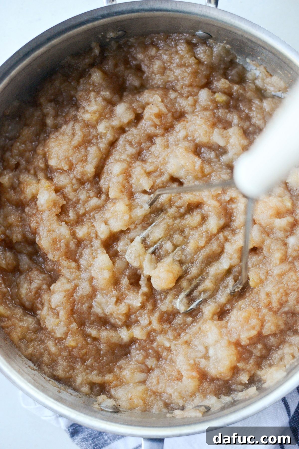 Mashing cooked apples in a skillet with a hand masher to create homemade applesauce.