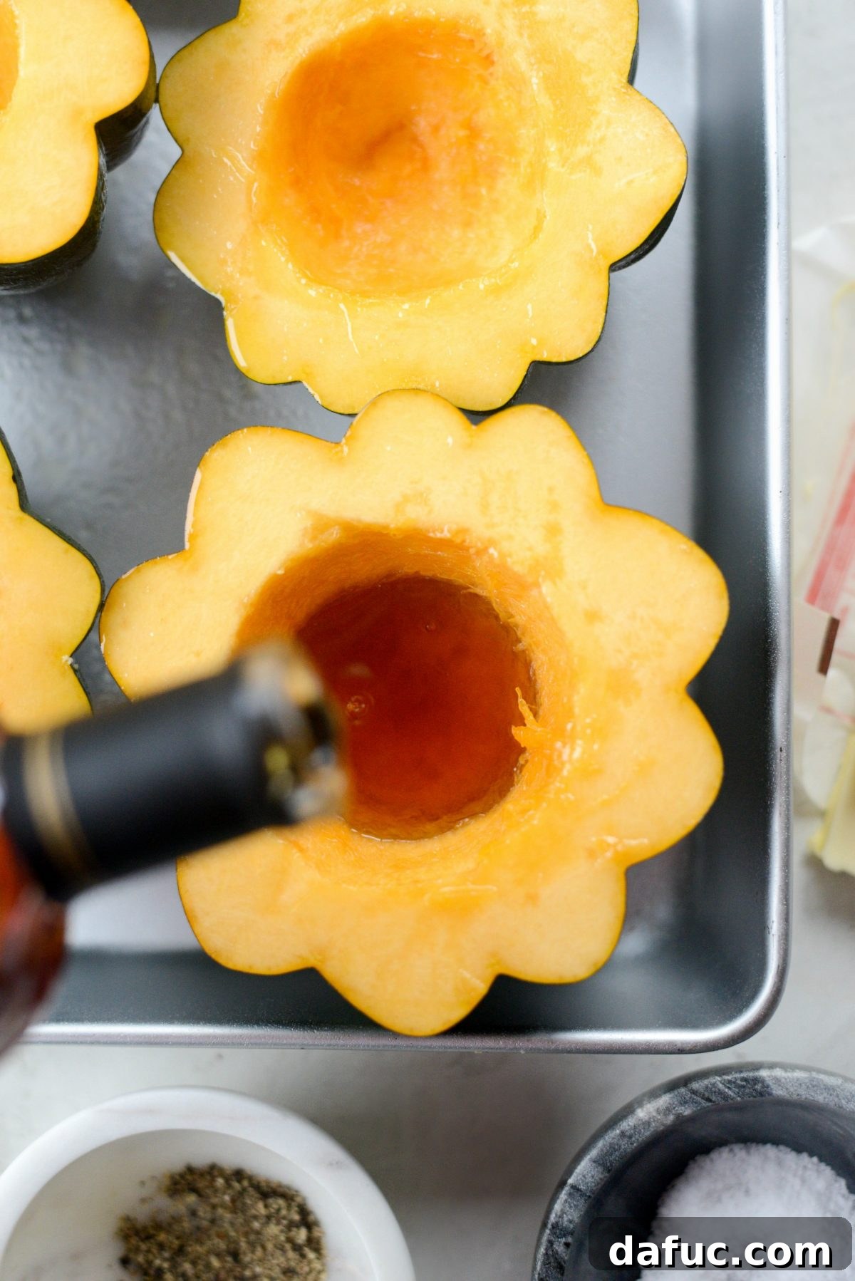 Maple syrup being poured generously into the cavity of an acorn squash half in a roasting pan, ready for roasting.