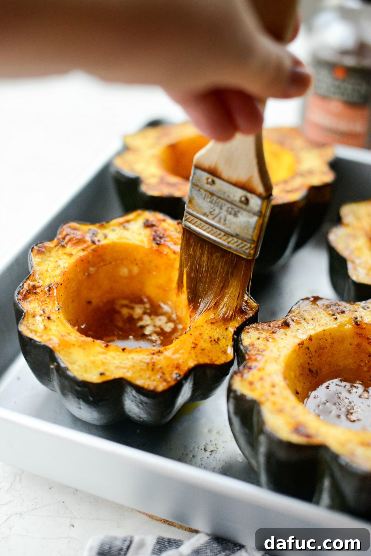 A pastry brush applying the caramelized maple butter glaze over the tender flesh of roasted acorn squash halves.