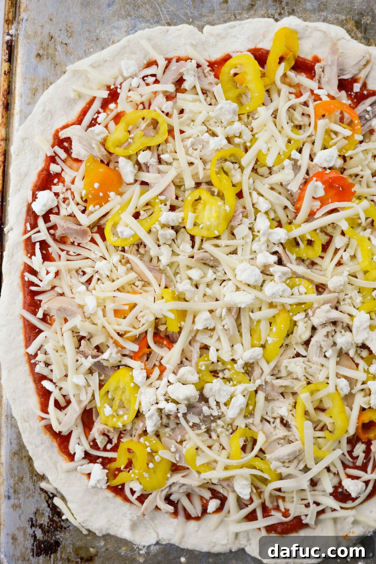 Topping the Spicy Chicken Feta Pizza with shredded chicken, banana peppers, mozzarella, and crumbled feta cheese before baking