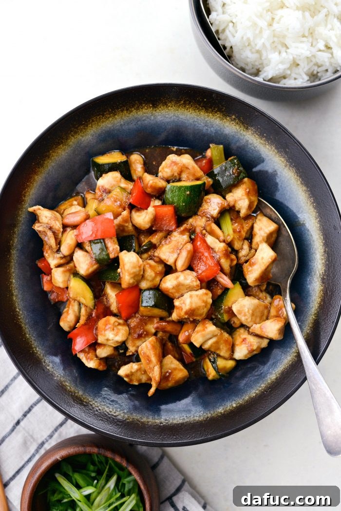 A elegant blue and black serving dish filled with a generous portion of Kung Pao chicken, alongside a small bowl of rice and a garnish of green onions, ready for a meal.