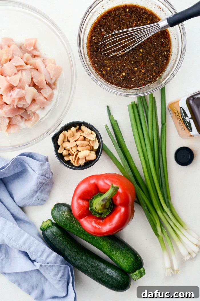 A selection of fresh ingredients for Kung Pao Chicken, including diced chicken, Kung Pao sauce, peanuts, green onions, red bell pepper, zucchini, and peanut oil, arranged on a rustic surface.