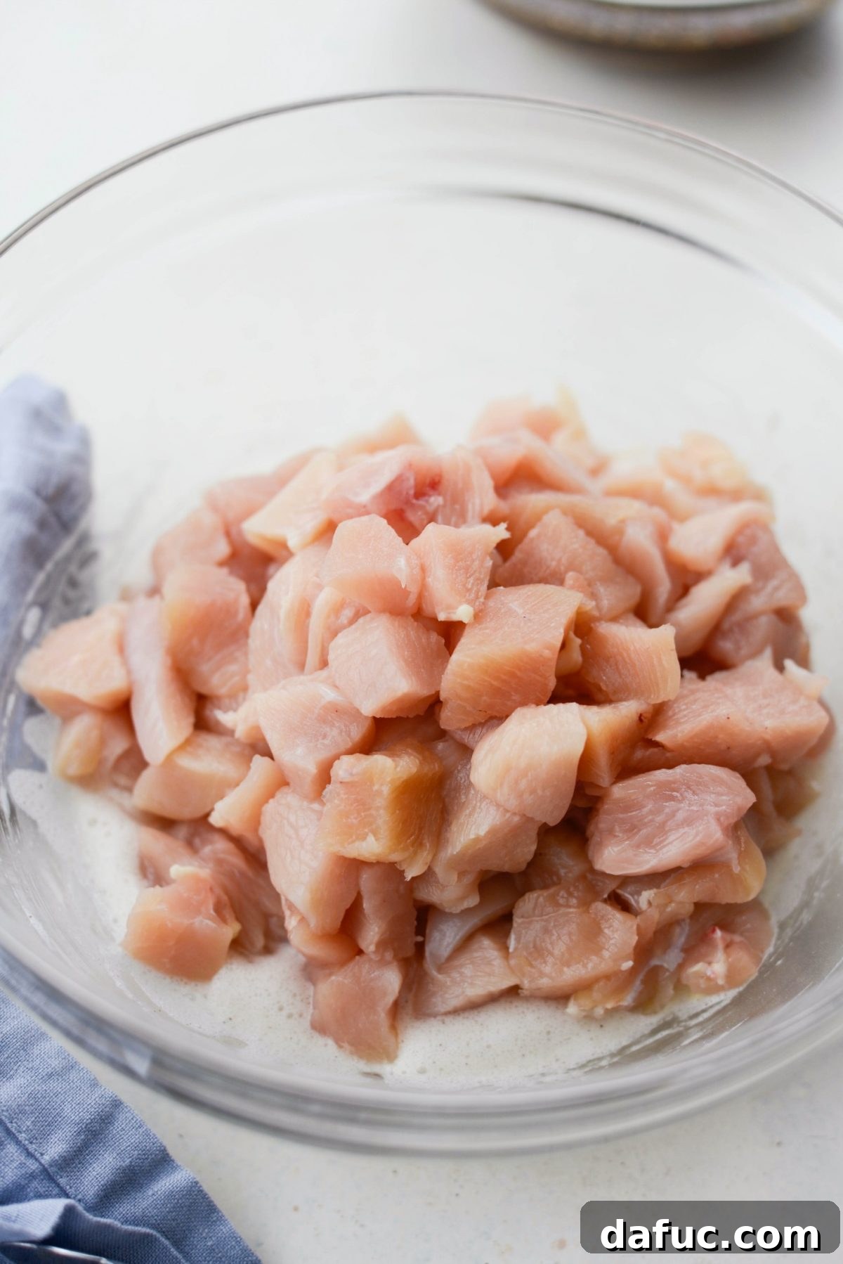 Diced chicken pieces in a glass bowl, coated with whipped egg whites, cornstarch, and white pepper, ready for cooking.