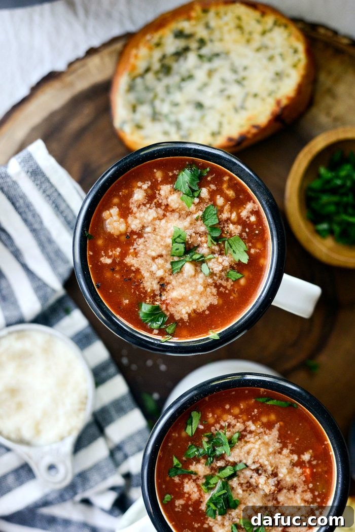 A steaming bowl of hearty Beef Tomato Ancini de Pepe Soup, garnished with fresh herbs and Parmesan, served on a rustic wooden surface. SimplyScratch.com