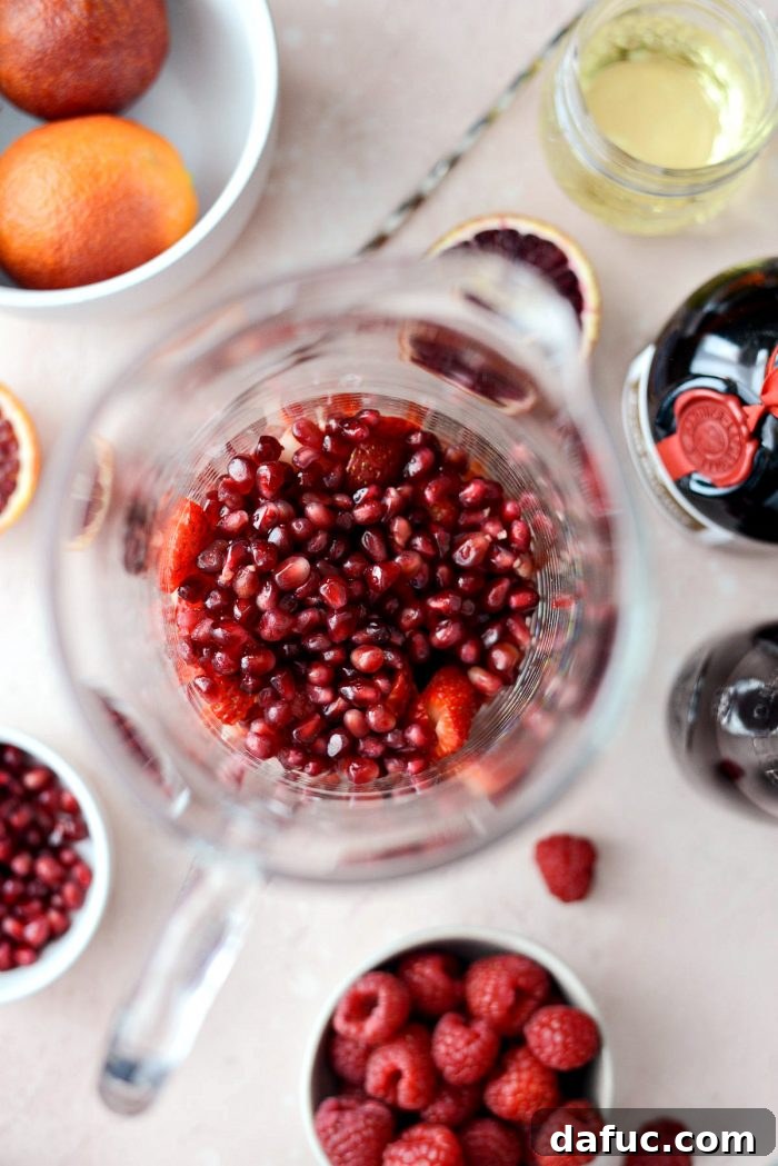 Preparing Champagne Sangria: Sliced strawberries and pomegranate arils are added to a large glass pitcher.