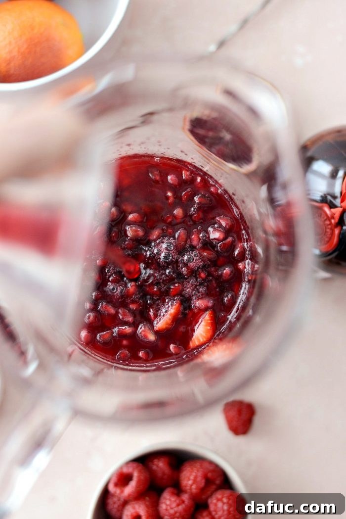 Pomegranate juice and freshly squeezed blood orange juice being poured into a pitcher with fruit for Champagne Sangria.