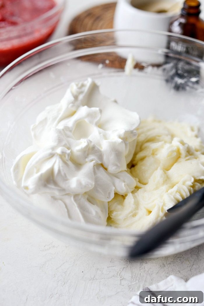 Unsweetened whipped cream being carefully folded into the sweetened cream cheese mixture with a rubber spatula.