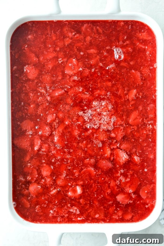 The chilled strawberry Jell-O mixture being poured carefully over the set cream cheese layer in the baking dish.