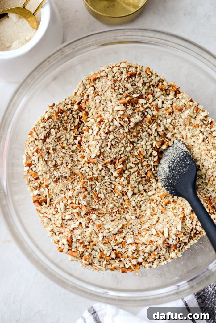 A clear mixing bowl filled with a mixture of pretzel crumbs, ground almonds, and sugar, ready for butter.
