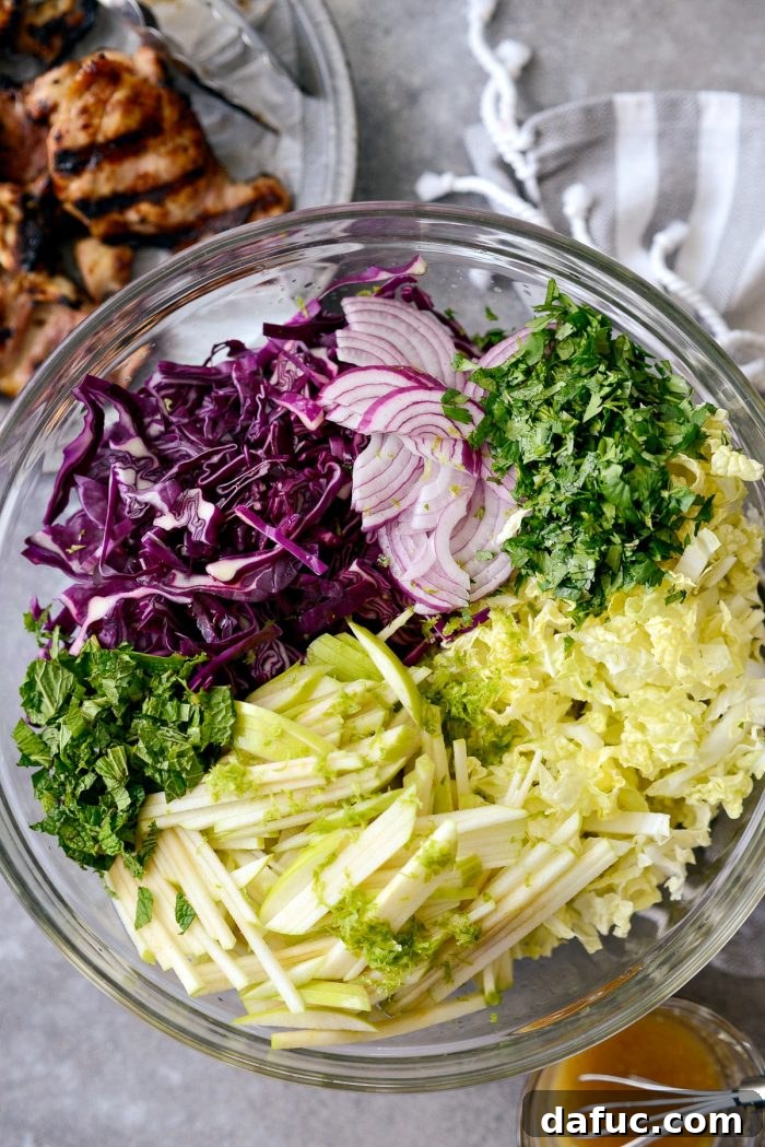 Vibrant Grilled Thai Chicken Salad 14 Large glass bowl filled with chopped purple and Napa cabbages, matchstick green apple, thinly sliced red onion, fresh mint, cilantro, lime zest, and lime juice.