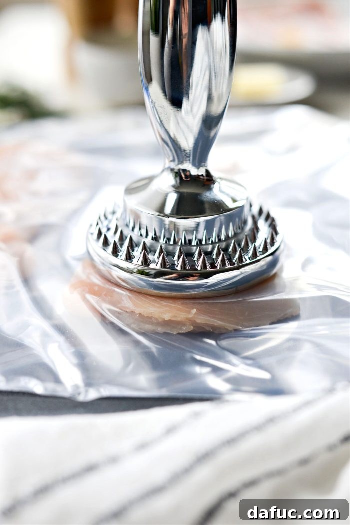 Signature Chipotle Chicken Marinade 5 A close-up shot of a raw boneless, skinless chicken breast being flattened to an even thickness. It is placed between two sheets of clear plastic wrap, with a flat meat tenderizer poised above it, illustrating the preparation process for even cooking.