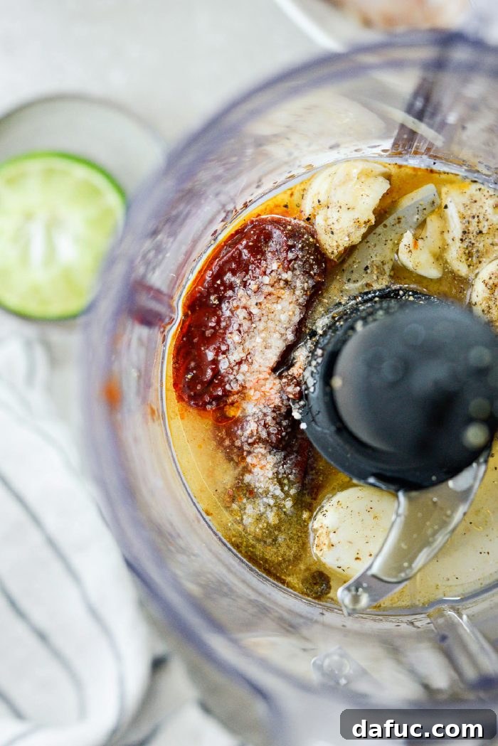 Signature Chipotle Chicken Marinade 6 All the marinade ingredients, including whole chipotle peppers, peeled garlic cloves, a wedge of fresh lime, a drizzle of honey, a bottle of avocado oil, and bowls of ground cumin, salt, and pepper, meticulously arranged inside a mini food processor bowl.