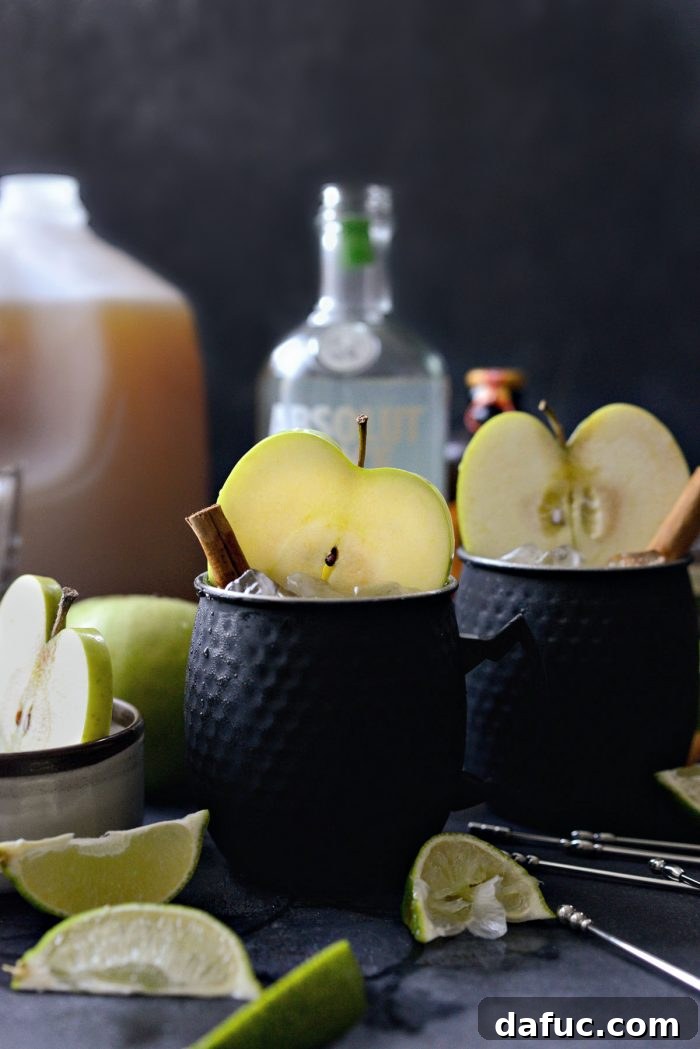 Orchard Mule 3 Two Apple Cider Moscow Mules served in sleek black metal cups, garnished with lime and apple slices, perfect for a modern autumn gathering.