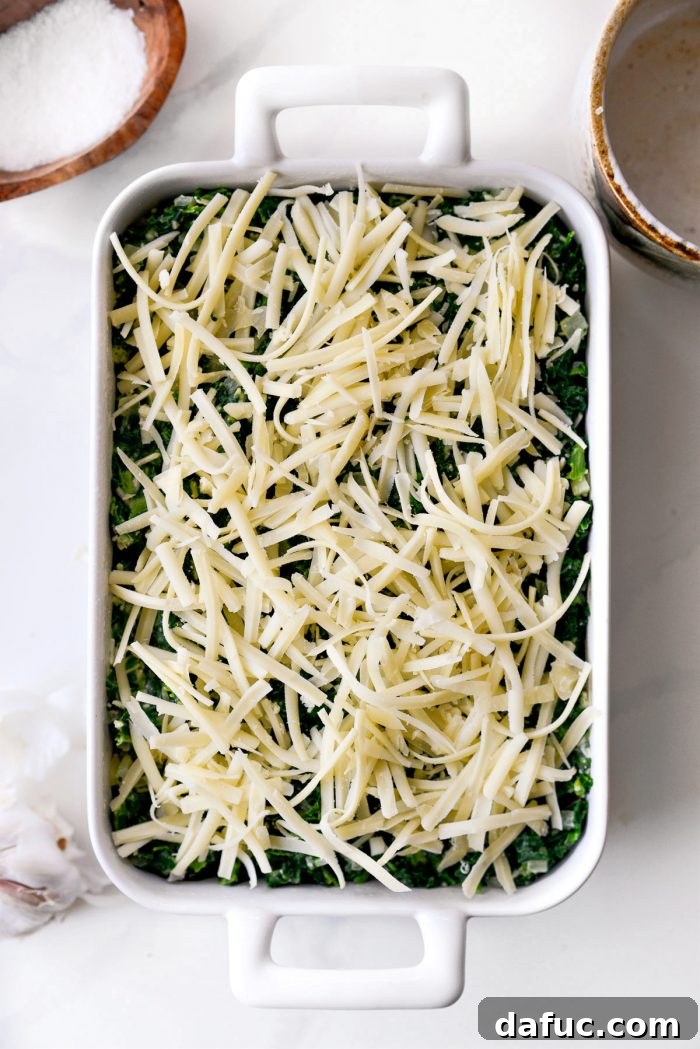 Grated Gruyere cheese being generously sprinkled over the spinach mixture in the casserole dish before baking.