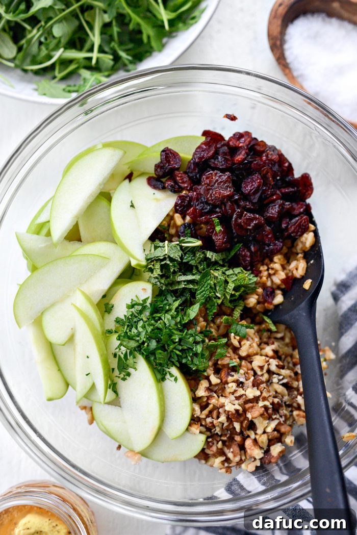 Cooked farro, sliced green apples, dried cranberries, and fresh herbs combined in a large mixing bowl, ready for the next additions.