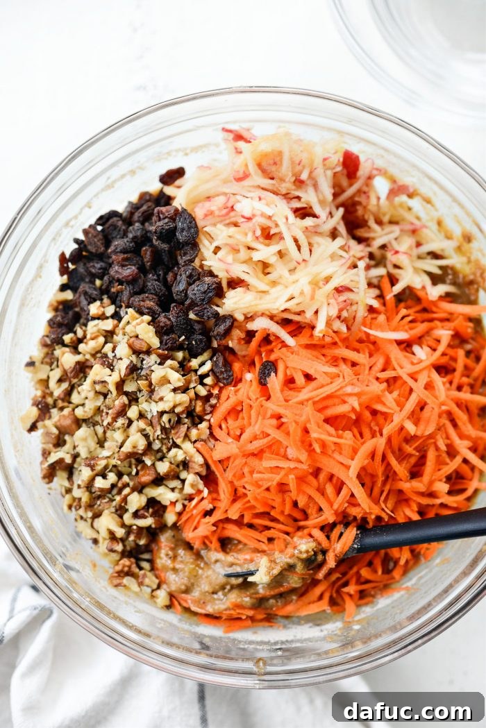 Shredded carrots, grated apples, raisins, and chopped walnuts being folded into muffin batter.