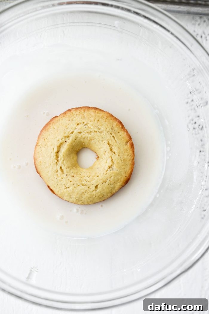 A Glazed Buttermilk Cake Doughnut being dipped halfway into the vanilla glaze.