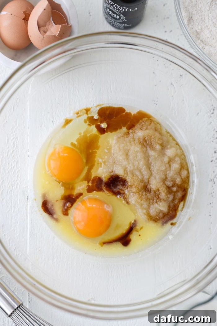 A glass mixing bowl containing wet ingredients for doughnuts: eggs, unsweetened applesauce, melted butter, and vanilla extract.
