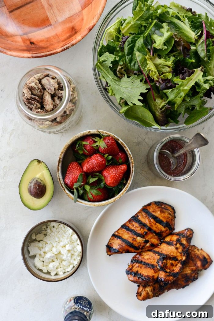 Various fresh ingredients laid out for Strawberry Goat Cheese Spring Salad preparation, including greens, strawberries, avocado, goat cheese, and dressing components