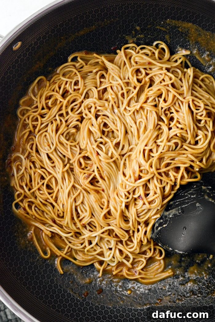 Fiery Garlic Sesame Noodles 15 Tongs actively tossing cooked noodles with the thick, glossy spicy garlic sesame sauce in a wok to ensure full coating.