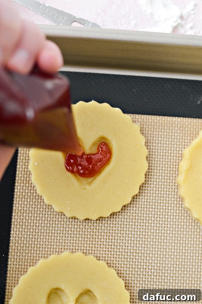 Jam Filled Butter Heart Cookies 14 Filling the heart-shaped indentations in raw cookie dough with vibrant raspberry jam using a piping bag, ensuring careful and even distribution.