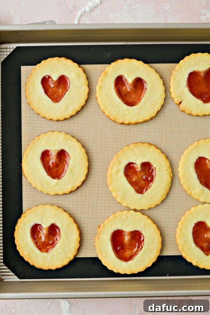 Jam Filled Butter Heart Cookies 16 Freshly baked Jam Heart Butter Cookies on a baking sheet, displaying perfectly golden edges and glistening, set jam centers.