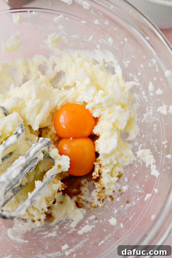 Jam Filled Butter Heart Cookies 7 Adding two bright yellow egg yolks, vanilla extract, and almond extract to the creamed butter and sugar mixture in the mixing bowl.
