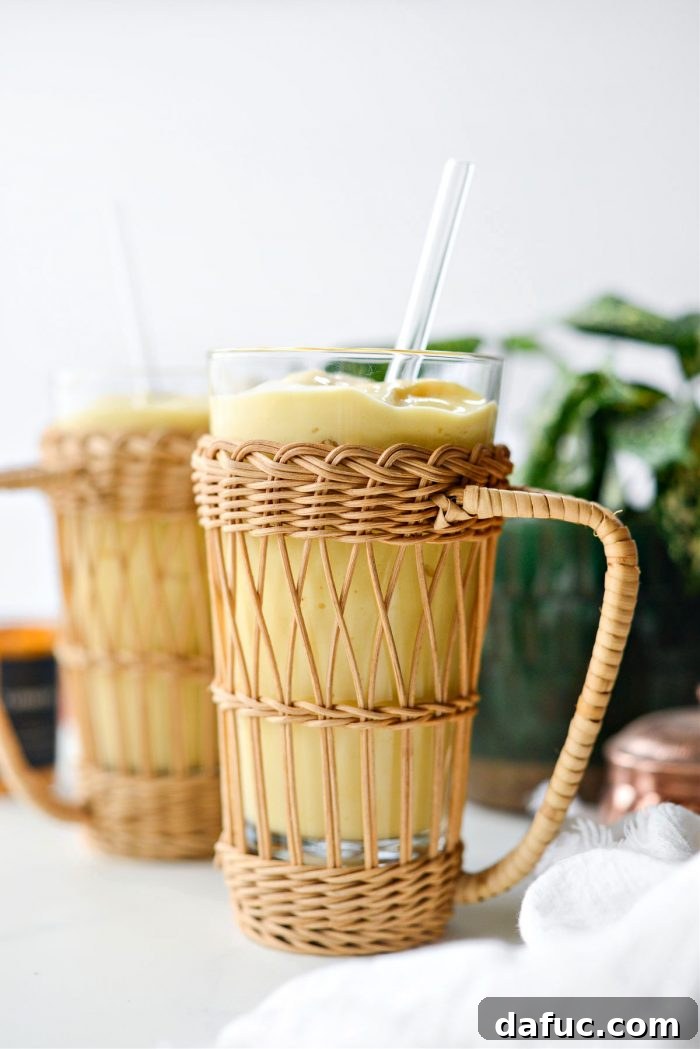 Liquid Sunshine Mango 2 Golden Hour Mango Smoothie, a vibrant yellow-orange smoothie in a glass with tropical fruit in the background.